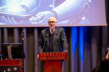 A person in a suit is speaking from a lectern with the inscription ‘Veritatis Splendor’, standing in front of a large screen displaying a futuristic graphic of a robot holding a tablet.