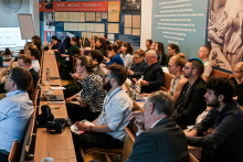 A group of people sitting in a room. They are dressed both formally and informally. They are looking ahead, appearing to be listening to something. Some people are taking notes. The walls of the room are covered with colorful informational boards.
