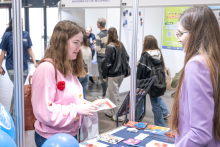 Two people are standing at the trade fair booth, one of them holding a leaflet. Informational materials are laid out on the table, and other participants in the exhibition hall can be seen in the background.