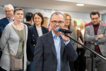 A person standing by a microphone during an indoor event, addressing the gathered participants visible in the background.