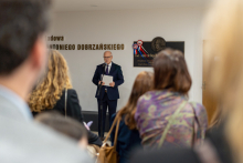 A man in a suit stands at a lectern and reads a text into the microphone, addressing the gathered audience; in the background, a commemorative plaque and the inscription 'Lecture Hall named after Prof. Antoni Dobrzański' are visible.