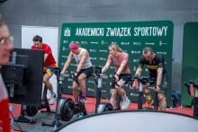 A woman and a man are exercising on stationary bikes arranged in a row during a sports event. In the background, a green wall with the logo of the Academic Sports Association is visible
