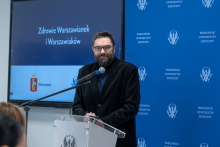 A person is standing at a lectern and giving a speech. They are resting their hands on the podium and looking toward the audience. A microphone is positioned next to them. In the background, a blue wall with the logo of the Medical University of Warsaw is visible. Next to it, there is a screen displaying a slide concerning the health of the residents of Warsaw.