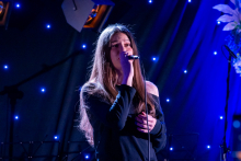 A woman is standing on stage and singing into a microphone. She is holding the microphone close to her mouth and tilting her head slightly. Her posture is calm, and her facial expression suggests she is focused on the performance. In the background, small lights are glowing, creating an atmospheric scene.