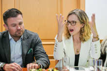 Two people are sitting at a table and talking. The woman on the left is wearing a grey blazer and a light blue shirt. The man on the right is wearing a white blazer and has his hands raised.