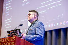 A person standing at a lectern with the inscription ‘Veritatis Splendor’, presenting content from a laptop decorated with colorful stickers. In the background, a large screen displaying an application interface is visible during the presentation.