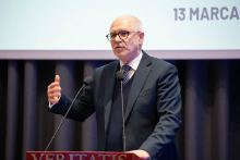 A person speaking at a podium during an event, gesturing with one hand, with part of a presentation slide showing the date ‘13 March’ in the background.