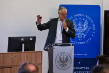 A middle‑aged man, dressed formally, is standing in front of a lectern. He is holding a microphone in his left hand, with his right hand raised. Behind him stands a blue banner with the WUM logo. Next to him, a desk with a computer is visible.