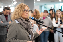 A person standing by a microphone during an indoor event, addressing the gathered participants visible in the background.