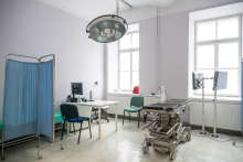 A bright hospital treatment room with a desk and computer, a privacy screen, a mobile treatment table on wheels, and a large surgical lamp mounted on the ceiling; in the background, two tall windows letting in natural light.