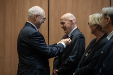 Rektor przypina odznaczenie innemu mężczyźnie podczas uroczystości. Obaj stoją naprzeciw siebie z poważnymi wyrazami twarzy. Kobieta i mężczyzna stojący obok spokojnie obserwują przebieg wydarzenia. Atmosfera jest uroczysta i skupiona.