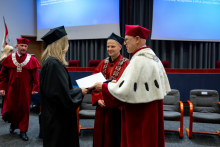 A woman in an academic gown stands opposite two professors and holds a document. The rector is shaking her hand, and another man stands beside him.