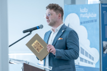 A person standing at a lectern, holding a book with a decorative cover, speaking into a microphone in an exhibition space.