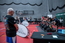 A man wearing a colourful T‑shirt with the Great Orchestra of Christmas Charity logo is standing in a sports hall and watching a group of women and men exercising on stationary bikes