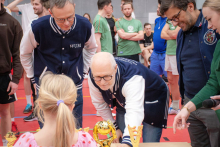 Osoby zgromadzone przy stole podczas dekoracji sportowej oglądają ustawione na nim puchary, a przedstawiciele organizatorów pochylają się nad nagrodami w hali sportowej.