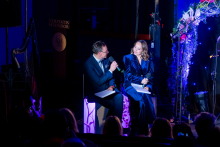 Two hosts are sitting next to each other and talking during the event. The man is holding a microphone and looking at the woman as if asking her a question. The woman is holding papers and responding, directing her gaze toward him. Their posture is relaxed, and their facial expressions suggest they are engaged in the conversation. The audience is seated in front of them in soft, dim lighting