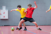 Two players in yellow and red jerseys are dynamically fighting for the ball during an indoor football match, blocking each other on the pitch.
