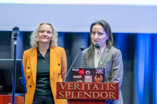 Two people are standing at a lectern with the inscription ‘Veritatis Splendor’, presenting content from a laptop decorated with colorful stickers. Next to them is a computer workstation, and in the background curtains and a projection screen are visible.