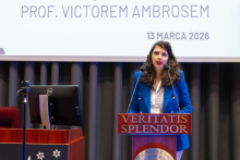 A person standing at a podium and speaking during an event, with a presentation slide behind them showing the date ‘13 March 2026’ and part of the session title.