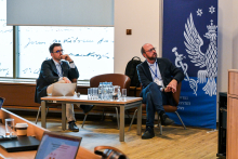 Two formally dressed men are sitting on chairs in the middle of the room, with a table visible between them. Their heads are turned to the right, as if they are listening to something. Behind them, a navy‑blue WUM banner with a white logo is visible.