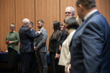 Eight people are taking part in the ceremony. The rector is pinning a decoration onto a man standing in front of him, who maintains a calm, focused expression. Several elegantly dressed people stand nearby, waiting for their turn. The woman on the left is holding a box with the decoration. The atmosphere is official and ceremonial.