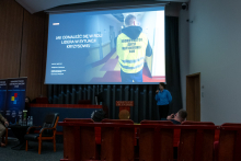 The speaker is standing with a microphone on the right side of the room, delivering a presentation. Behind her, a slide is displayed with the title of the presentation about the role of a leader in a crisis situation and a photo of a person wearing a yellow vest. Participants of the event are seated in the front rows.