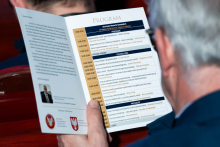 A person is seated in the auditorium, holding an open event program in their hands. The program has two pages and contains a detailed schedule of presentations with times and speakers’ names. The person bends their head toward the brochure and reads it attentively.
