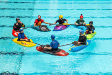 A group of people wearing helmets is sitting in kayaks arranged in a circle in a swimming pool. The participants are holding their paddles on the water, as if during a group exercise or kayaking instruction.