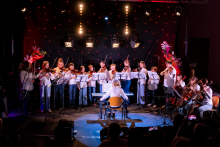 A group of a dozen young musicians is standing on stage, playing violins and cellos. They are all looking at the sheet music placed in front of them. In front of the ensemble, a conductor is seated, playing the piano and directing the performance. The audience is watching the show in dim lighting.