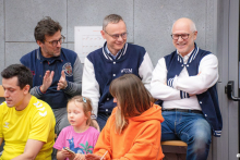 A group of people is sitting and standing by the wall in the sports hall; some of them are wearing navy blue jackets with white sleeves, while in front of them children and adults in colorful outfits are seated.