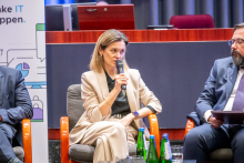 A person in a beige suit is sitting in an armchair and holding a microphone during a panel discussion. Next to them, other participants are seated, and bottles of water are placed on the table.
