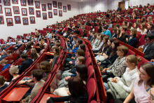 Audience seated in a large lecture hall, filling rows of red chairs; numerous framed portraits are visible on the left wall.