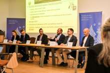 A group of people sitting at a table. They are dressed formally. On the table, there are white name cards with their first and last names. One of the individuals, a man, is holding a microphone and speaking into it. Behind them, a multimedia screen displaying a presentation is visible. There are also blue WUM banners with a white eagle.