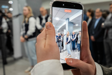 A shot of a person recording the event with a phone — the screen shows a scene with a speaker and a group of participants standing in the background in the lobby area.
