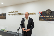 A man standing at a microphone is speaking, holding a phone and a document in his hands; in the background, a commemorative plaque with a medallion and the inscription 'Lecture Hall named after Prof. Antoni Dobrzański' are visible