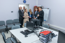 A group of people is standing at the entrance to a room equipped with desks with computers and chairs; one person is explaining something while standing closer to the board and the desks.