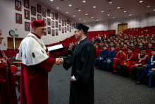 A man in a black academic gown receives a diploma from the rector. In the background, other people are seated on red chairs.