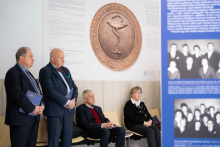 People standing and sitting in an exhibition space beneath a large bas‑relief featuring the emblem of the Faculty of Pharmacy, next to boards displaying information and archival photographs.