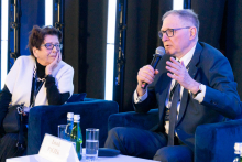Two people are sitting next to each other during a panel discussion. One person is holding a microphone and making a hand gesture, while the other is listening with their head resting on their hand. In front of them is a small table with water bottles and glasses.