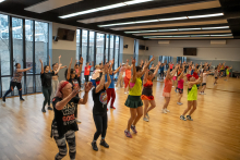 A group of people is participating in a dance class in a large fitness studio. They are all standing in several rows and raising their arms, performing the same movement. The room has a wooden floor, mirrors on the walls, and large windows with a view outside.