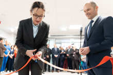 A person cutting a red ribbon during an indoor ribbon‑cutting ceremony, with another formally dressed person standing nearby and a group of attendees gathered in the background.
