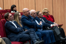 Six people are sitting in a single row of red seats, listening to the presentation. They are resting their hands on their knees or folding them in front of them. On the left sit a man and a woman, next to them three men, and at the end a woman dressed in red. More people are visible in the rows behind.