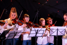 Six children are standing close together and playing the violin. Each of them is holding the instrument under their chin and looking at the sheet music placed on the stands.