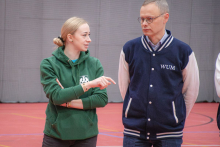 Two people are standing on the court in the sports hall; one, wearing a green sweatshirt, is pointing something out to the other person, who is dressed in a navy-blue jacket with white sleeves.