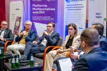 The panelists are seated in a semicircle on stage during the discussion, holding microphones. In the background, there are roll‑ups promoting IT solutions, and bottles of water are placed on the table in front of them.