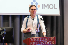 A person standing at a podium and speaking during a presentation, with the IMOL logo visible on the screen behind them