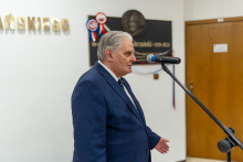 A man in a navy-blue suit stands at a microphone and gestures while speaking; in the background, a commemorative plaque with a medallion and a fragment of the inscription on the wall are visible.