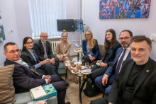 A group of people is sitting close together in a small room at a table with coffee, tea, and cookies, having a conversation in an informal atmosphere