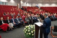 A speaker giving an address from a lectern during a ceremony in a large auditorium; in the front rows sit invited guests, and in the background there are filled red seats and a decoration made of white and yellow flowers.