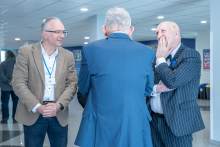 Three people standing in an exhibition hall, engaged in conversation and dressed in formal jackets.